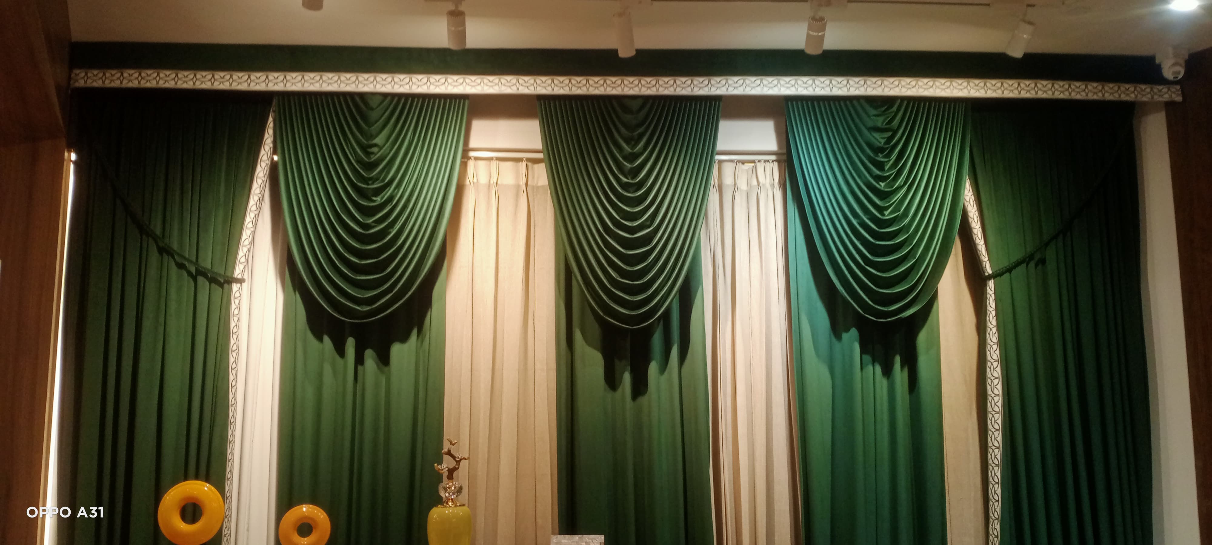 Grand theatrical emerald green swag valances draped over premium beige silk curtains on a showroom display wall